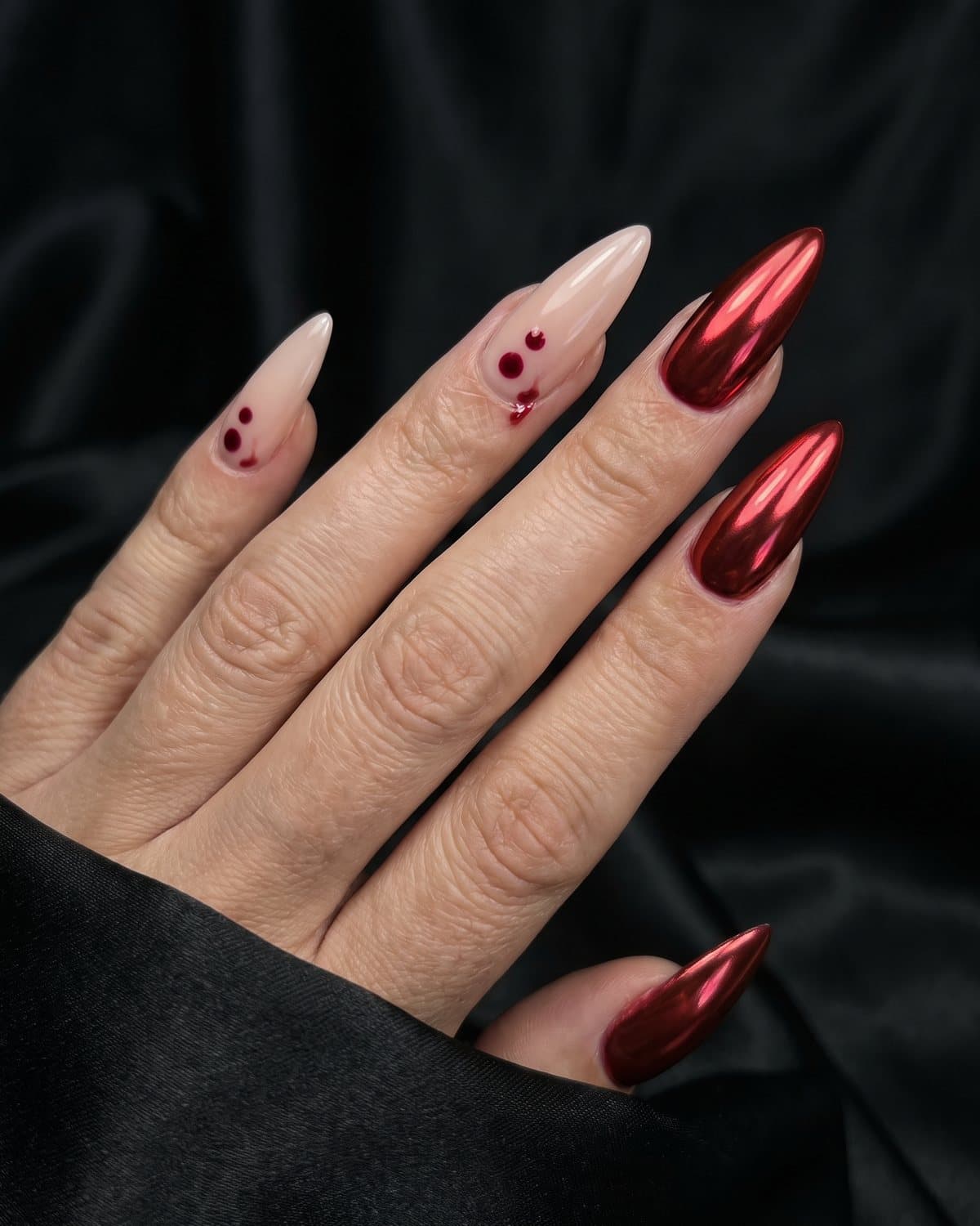 Stiletto nails with pale nude and two tiny red bite-mark dots with a small drip, and metallic red chrome nails, on black satin