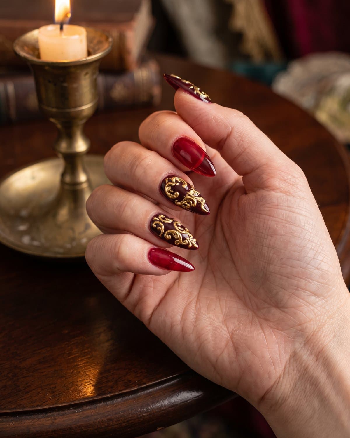 Stiletto nails in translucent deep red jelly and burgundy with ornate gold baroque filigree, beside an antique gold candlestick