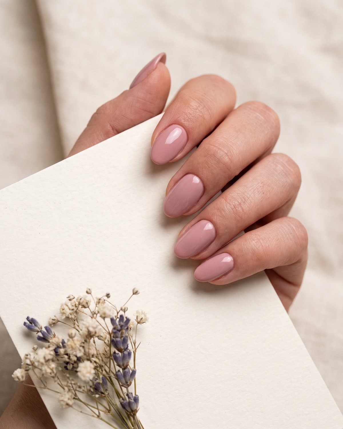 Glossy oval nails in soft dusty rose holding a blank cream wedding invitation with a dried flower