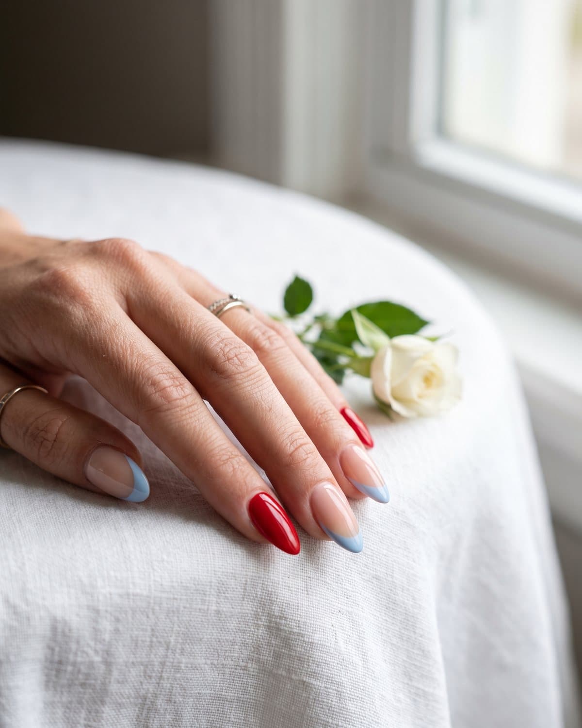 Almond nails with classic glossy red alternating with sheer nude nails with baby blue French tips, on a white tablecloth beside a white rose