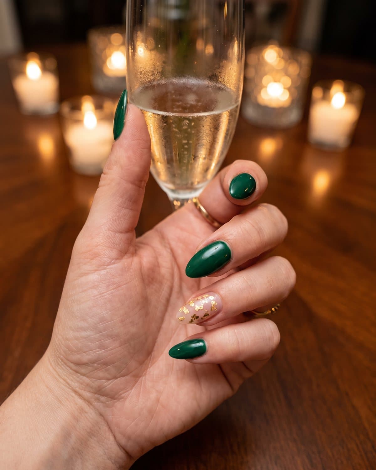 Almond nails in deep glossy emerald green with a gold foil accent nail, holding a champagne flute by candlelight