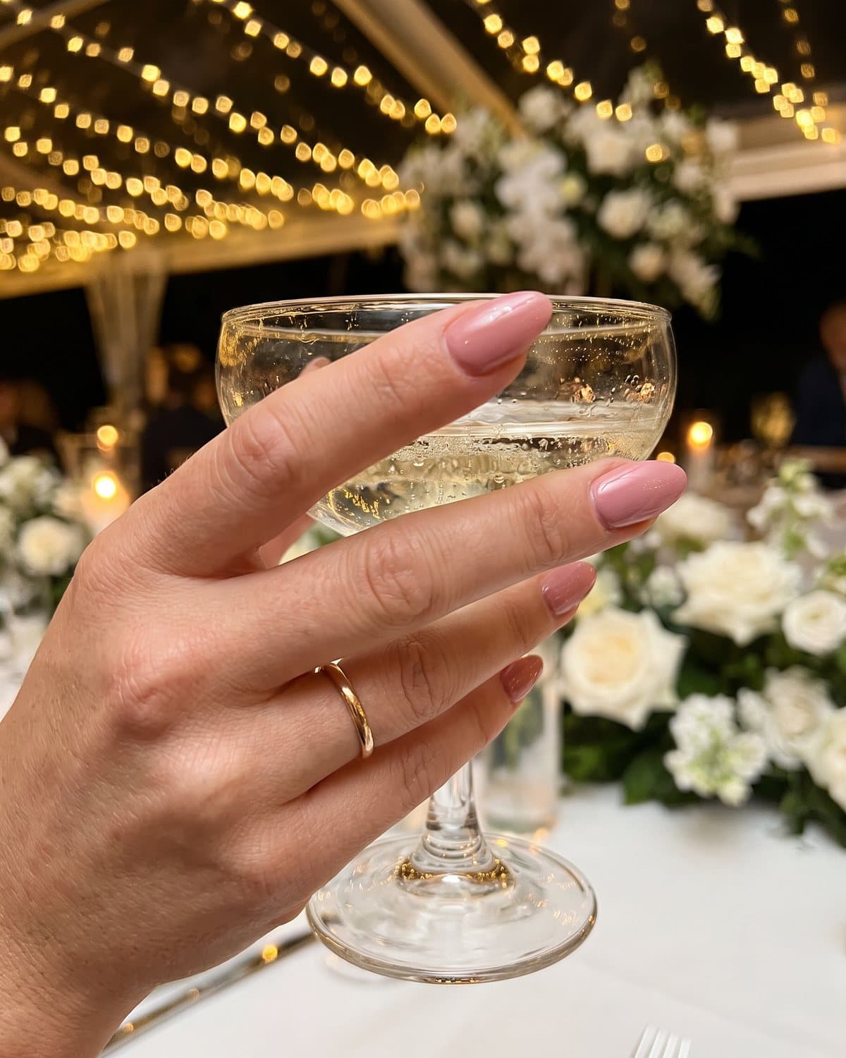 Glossy almond nails in soft dusty rose holding a champagne coupe at a wedding reception with string lights and white flowers