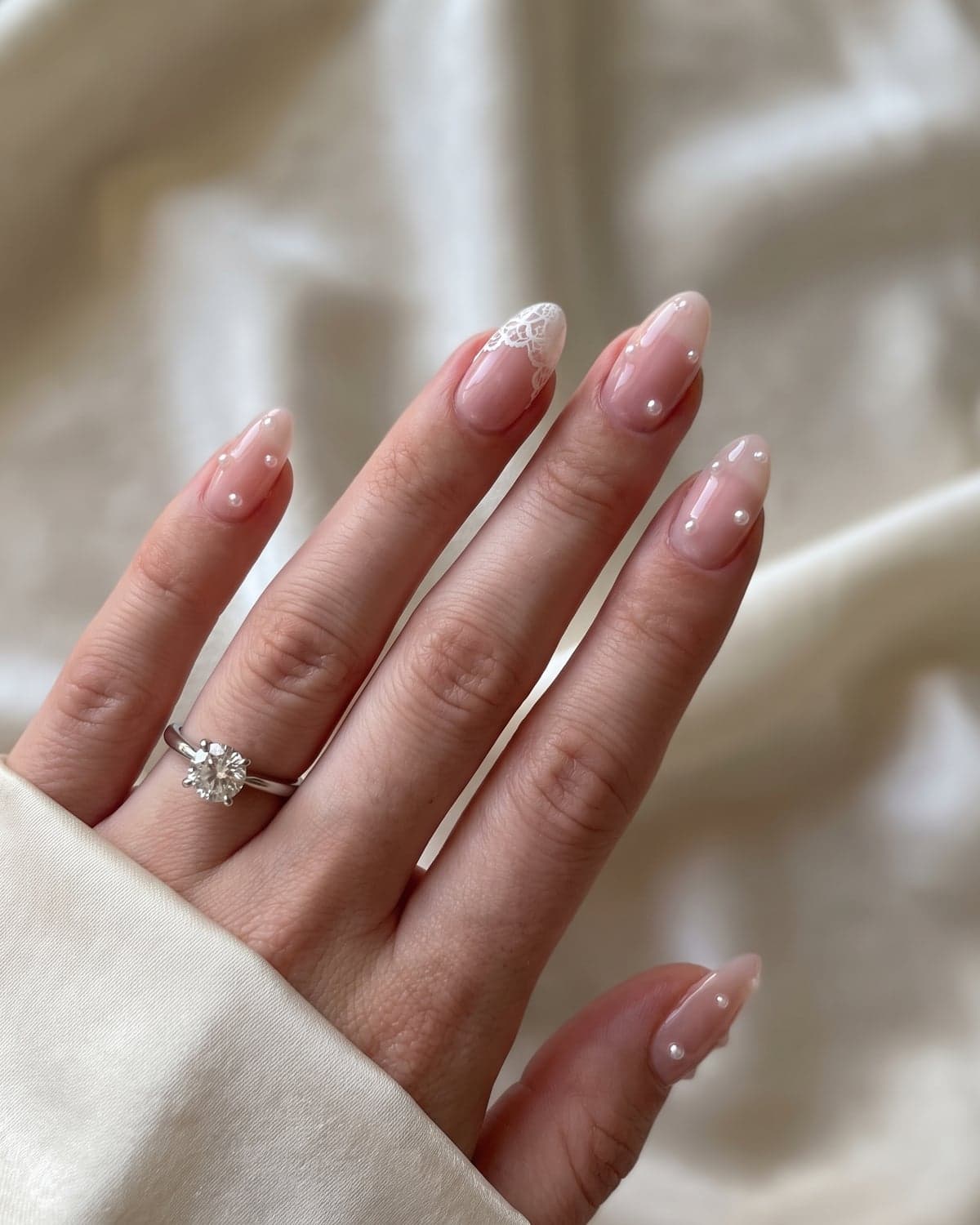 Sheer pink almond wedding nails with pearl accents and a white lace tip on an ivory background