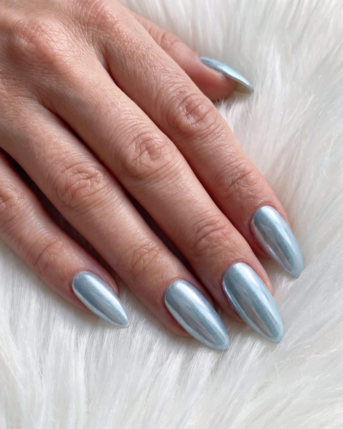 Almond nails in icy pale blue chrome with a frosty metallic sheen, resting on white faux fur