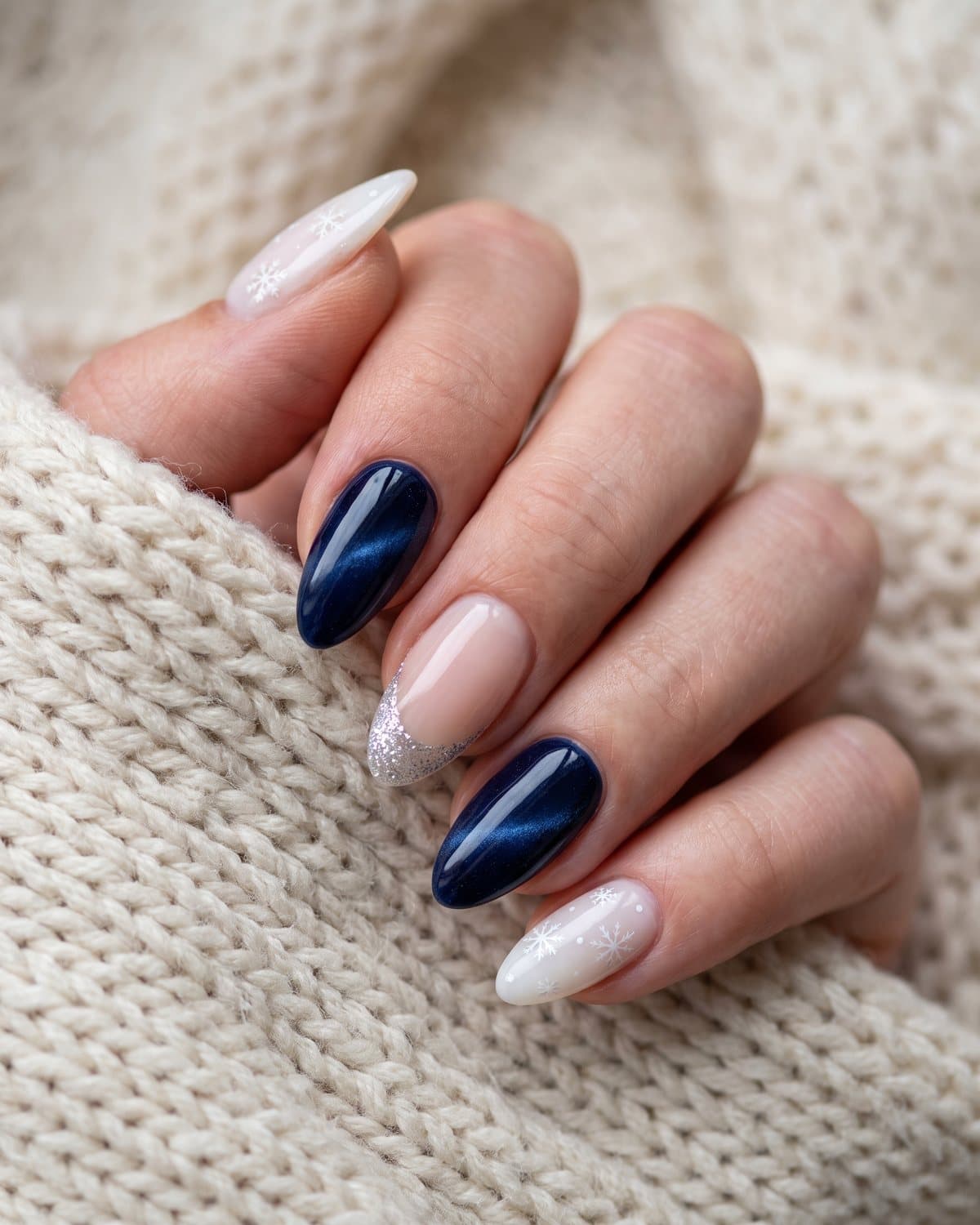 Almond nails with white snowflakes on milky white, deep navy velvet cat-eye and sheer nude with silver glitter French tips, on a cream knit scarf