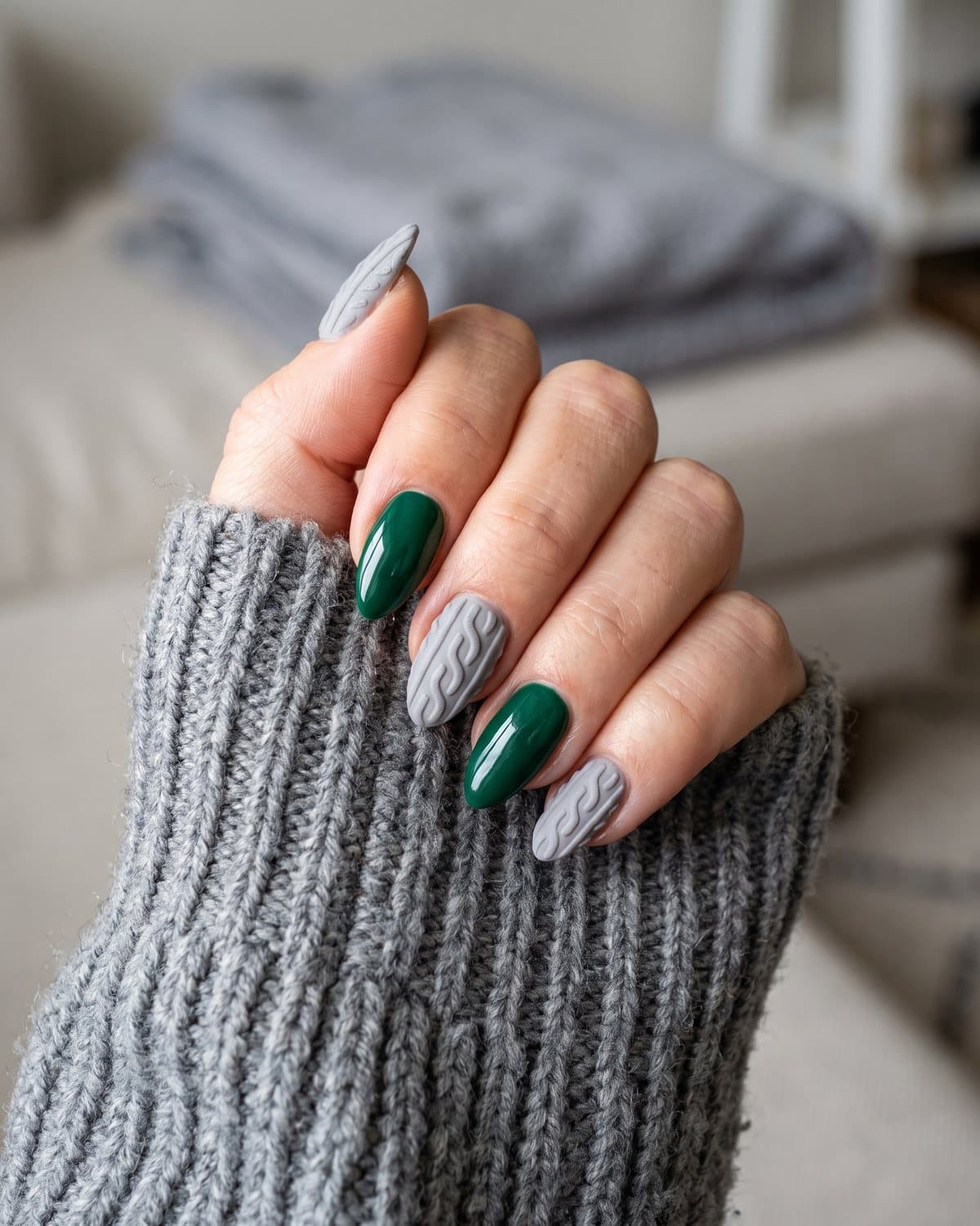 Almond nails with soft grey 3D cable-knit sweater texture and glossy deep emerald green, tucked into a grey chunky knit sleeve