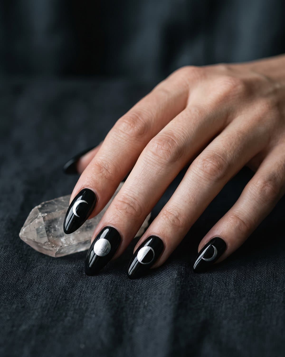 Glossy black almond nails with silver moon phases from crescent to full moon, resting on dark charcoal linen beside a crystal