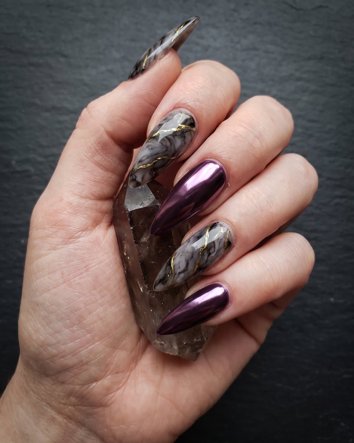 Stiletto nails with smoky grey and black quartz marble with gold veins alternating with deep plum chrome, holding a smoky quartz crystal