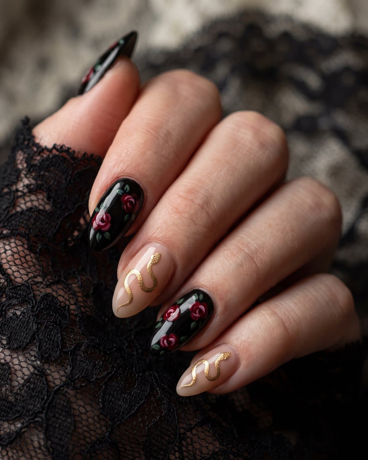 Almond nails with glossy black and small deep red roses alongside nude nails with a fine gold serpent, resting on black lace