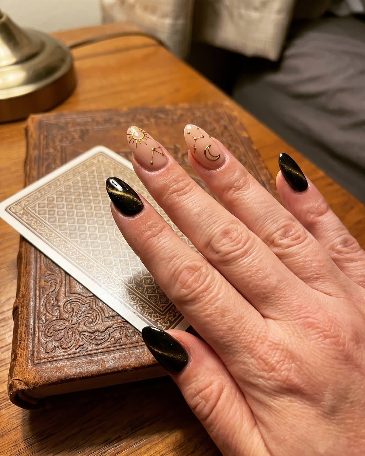 Almond nude nails with fine gold tarot-inspired suns and crescent moons alternating with black cat-eye nails with a gold shimmer line