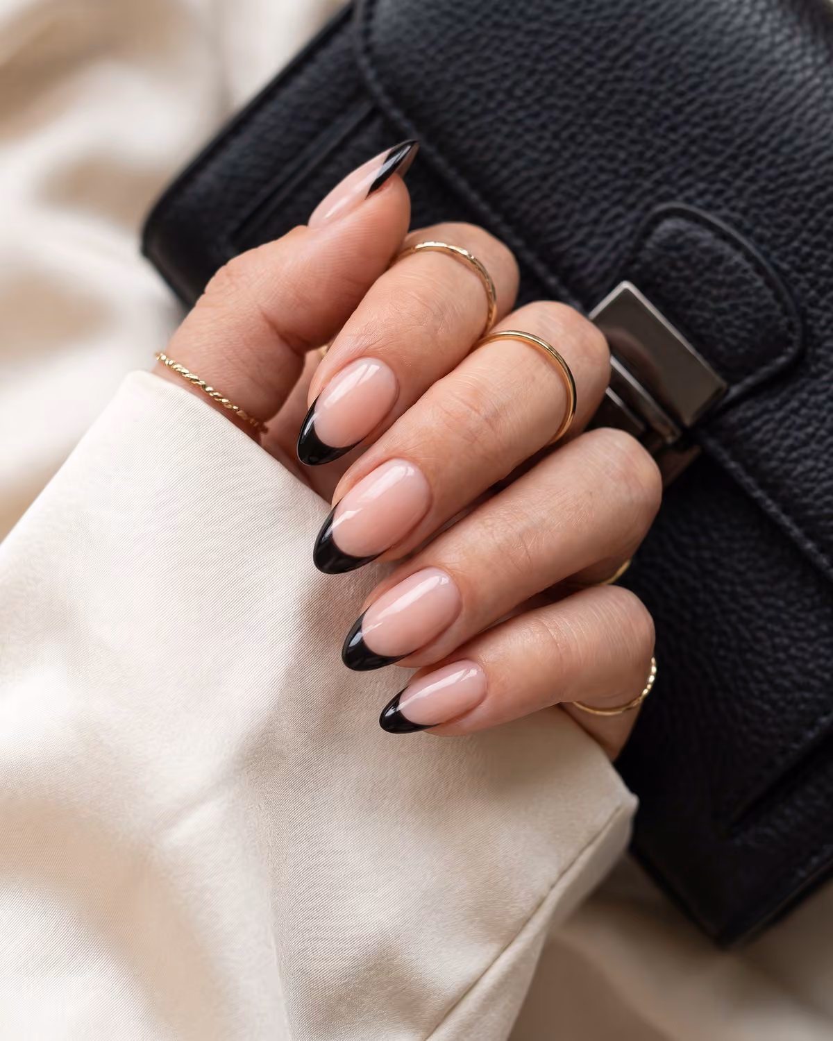 Glossy almond nails with a sheer pink nude base and crisp black French tips, next to a black leather handbag