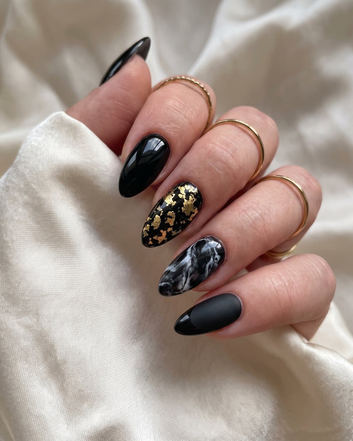 Almond black nail designs in glossy black, gold foil, black and white marble and matte black with glossy tips, with gold rings on cream silk