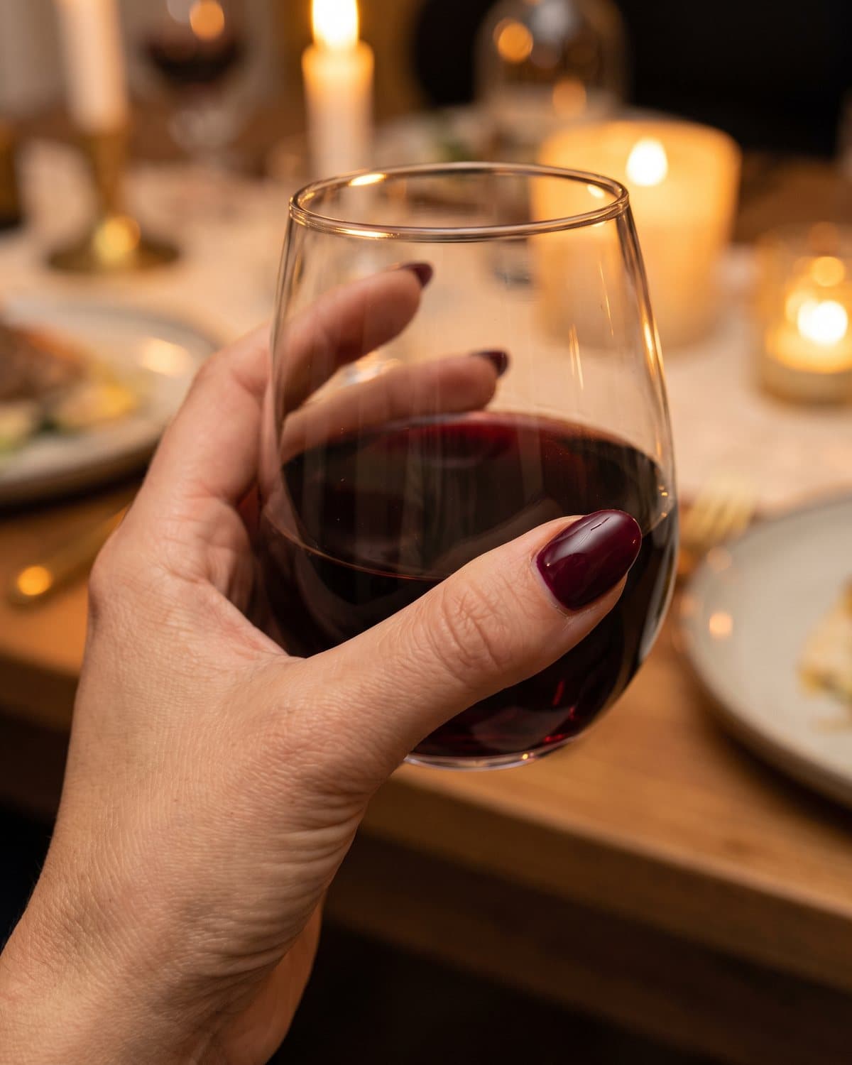 Glossy oval nails in deep burgundy holding a glass of red wine at a candlelit dinner