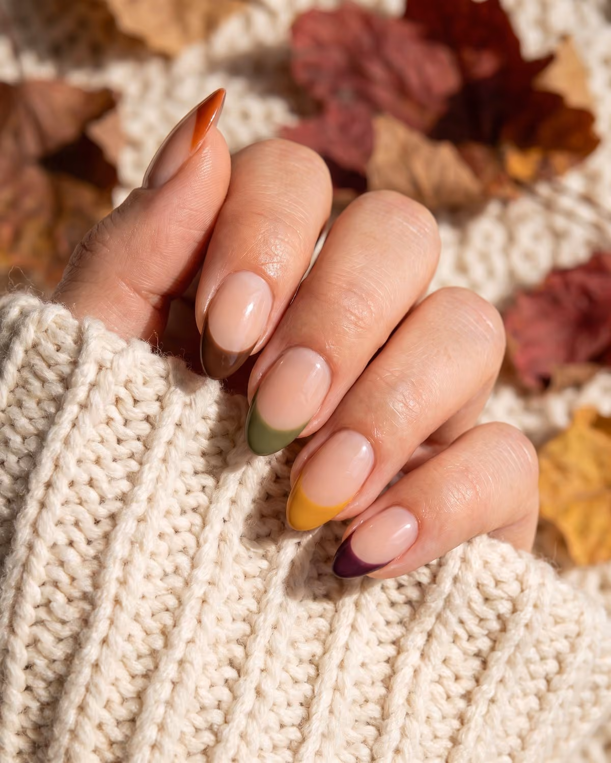 Almond nails with a sheer nude base and mismatched autumn French tips in burnt orange, chocolate, olive, mustard and plum among dried leaves