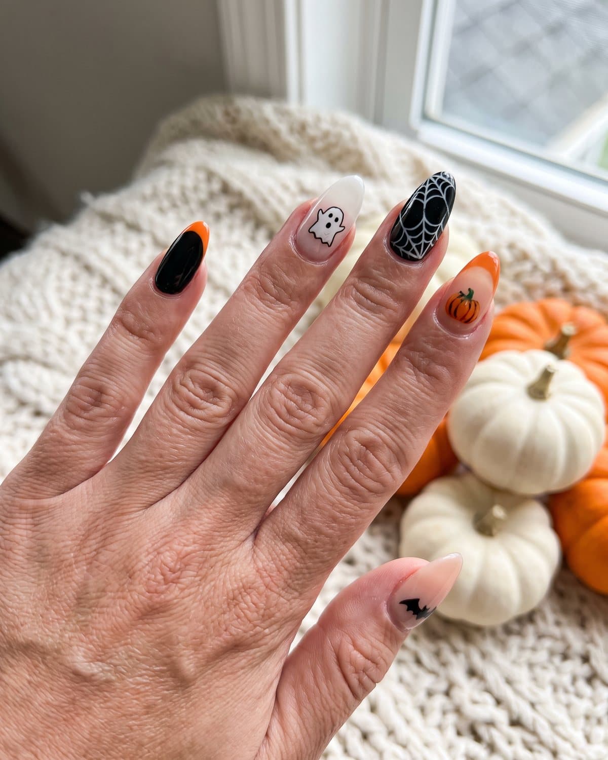Almond Halloween nails with an orange French tip, a tiny ghost, a spider web and a pumpkin, next to mini pumpkins on a cream knit blanket