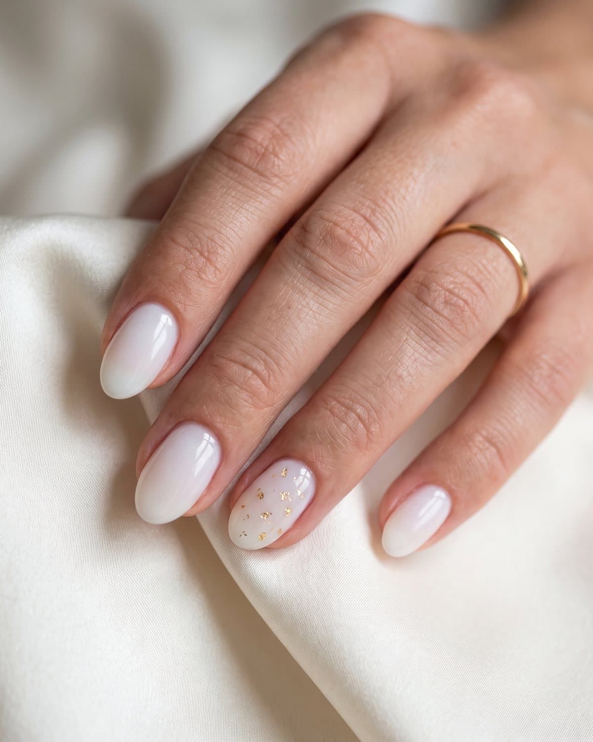 Glossy oval nails in luminous milky white with tiny gold flakes on one nail, resting on ivory silk with a gold ring