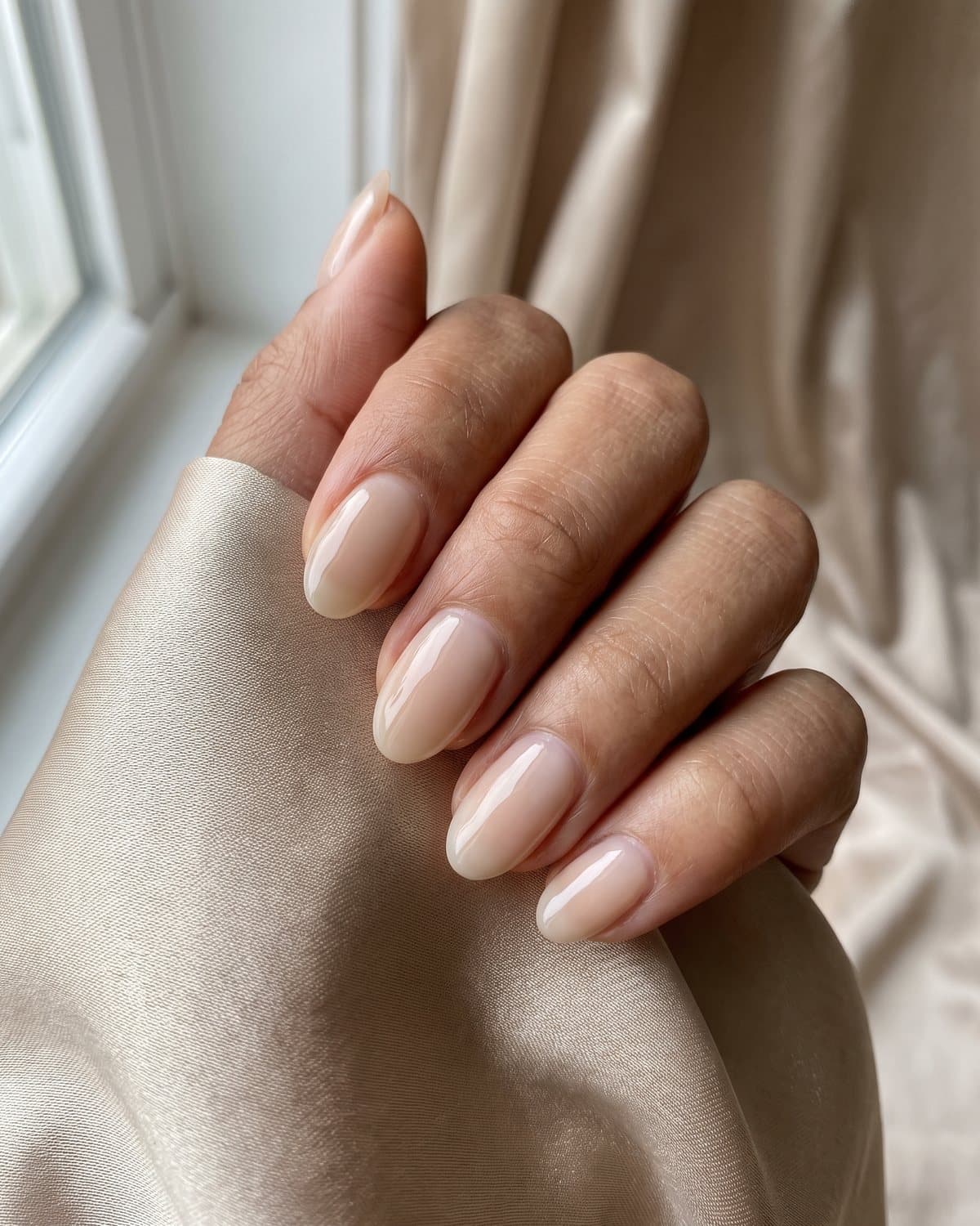 Medium skin tone hand with glossy oval nails in a milky beige nude, resting on draped champagne silk