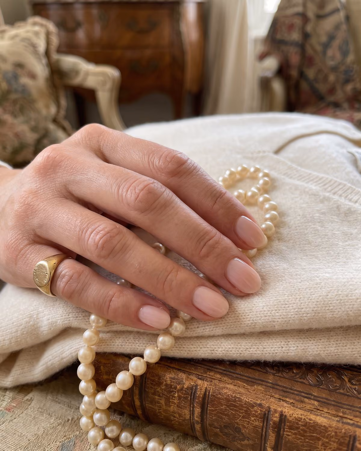 Short oval nails in sheer ballet pink with a gold signet ring, holding pearls on a cream cashmere sweater and leather-bound book