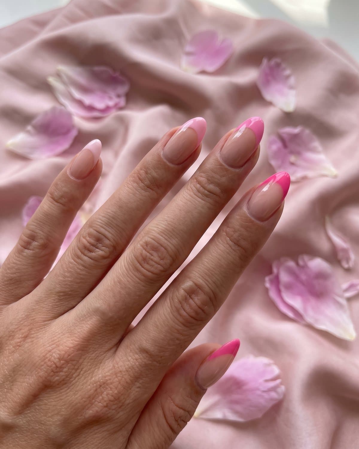Almond nails with a sheer nude base and pink French tips in shades from baby pink to hot pink, on blush satin with peony petals