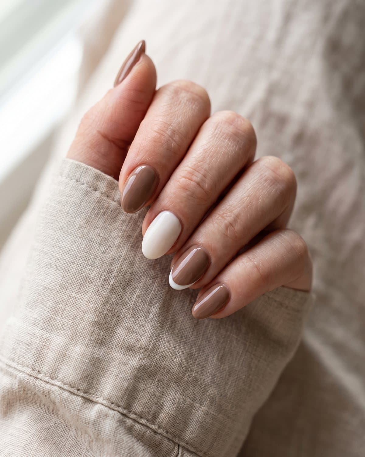 Short almond nails in glossy mocha brown and milky white with a micro French tip, resting on a beige linen shirt