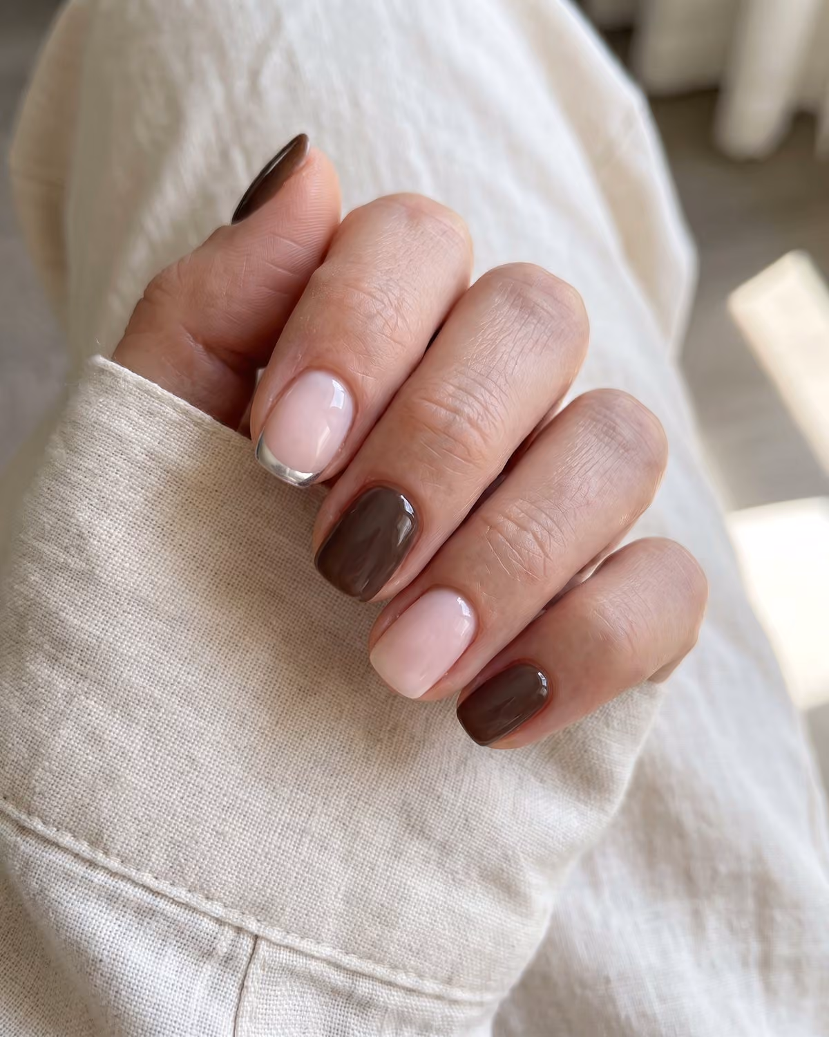 Short squoval gel nails in glossy espresso brown and milky pink with a silver chrome micro French tip, on a cream linen shirt