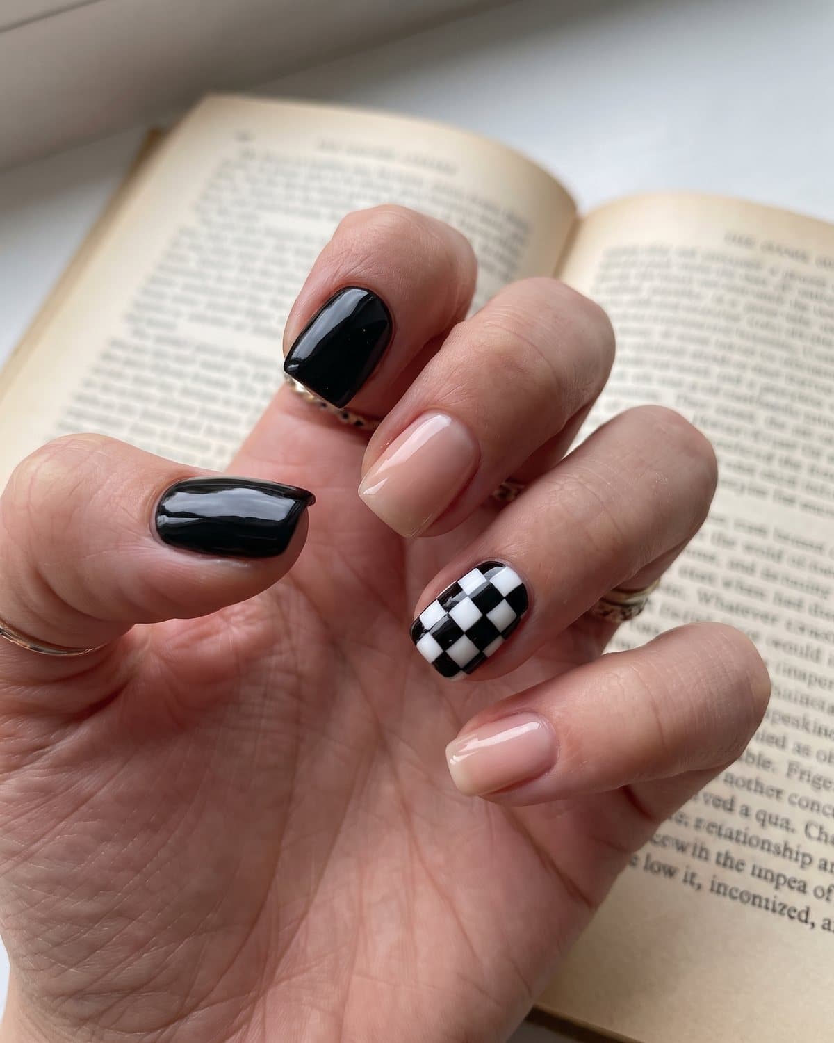 Short square nails with a black and white checkerboard accent, glossy black and sheer nude, resting on an open book