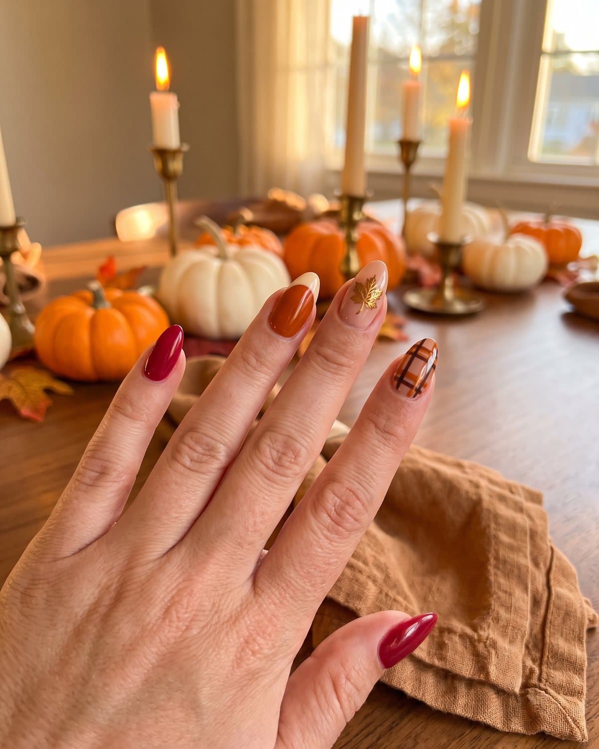 Almond Thanksgiving nails in cranberry, burnt orange French tip, gold leaf maple leaf and plaid on a table set with mini pumpkins and candles
