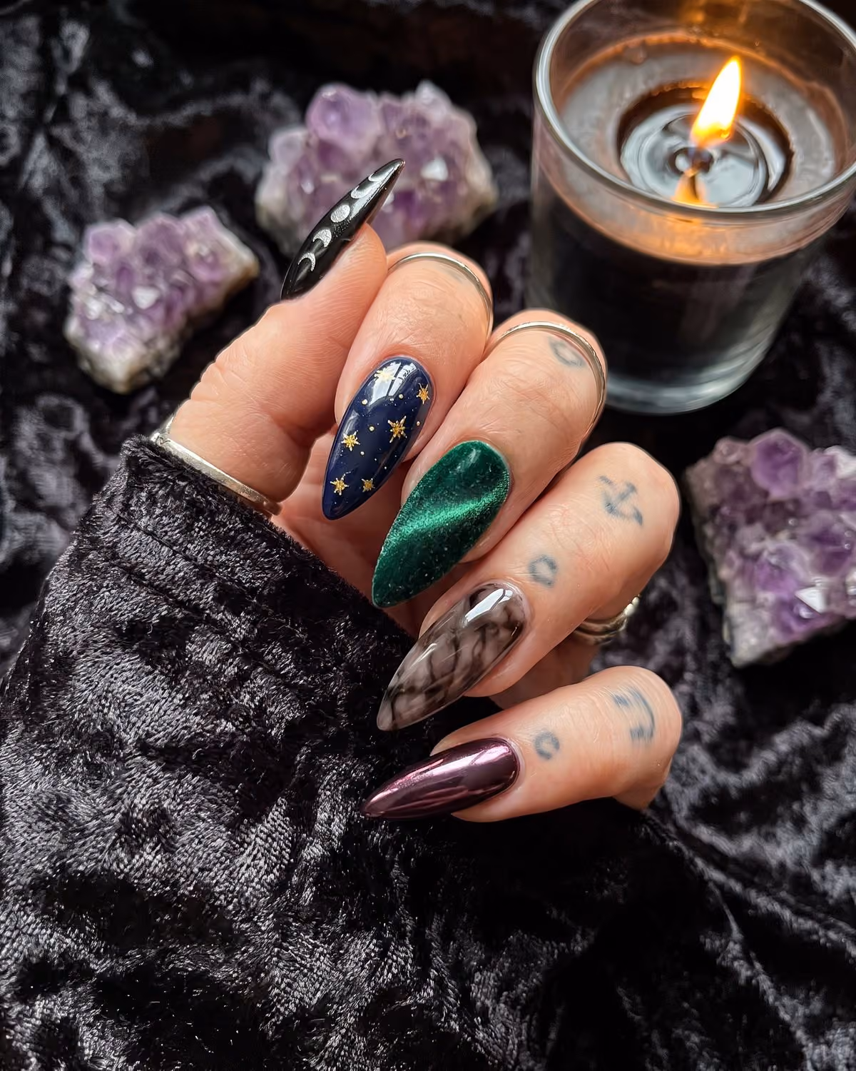 Witchy stiletto nails with silver moon phases, gold stars on navy, emerald velvet, smoky quartz and plum chrome, beside amethyst crystals and a candle